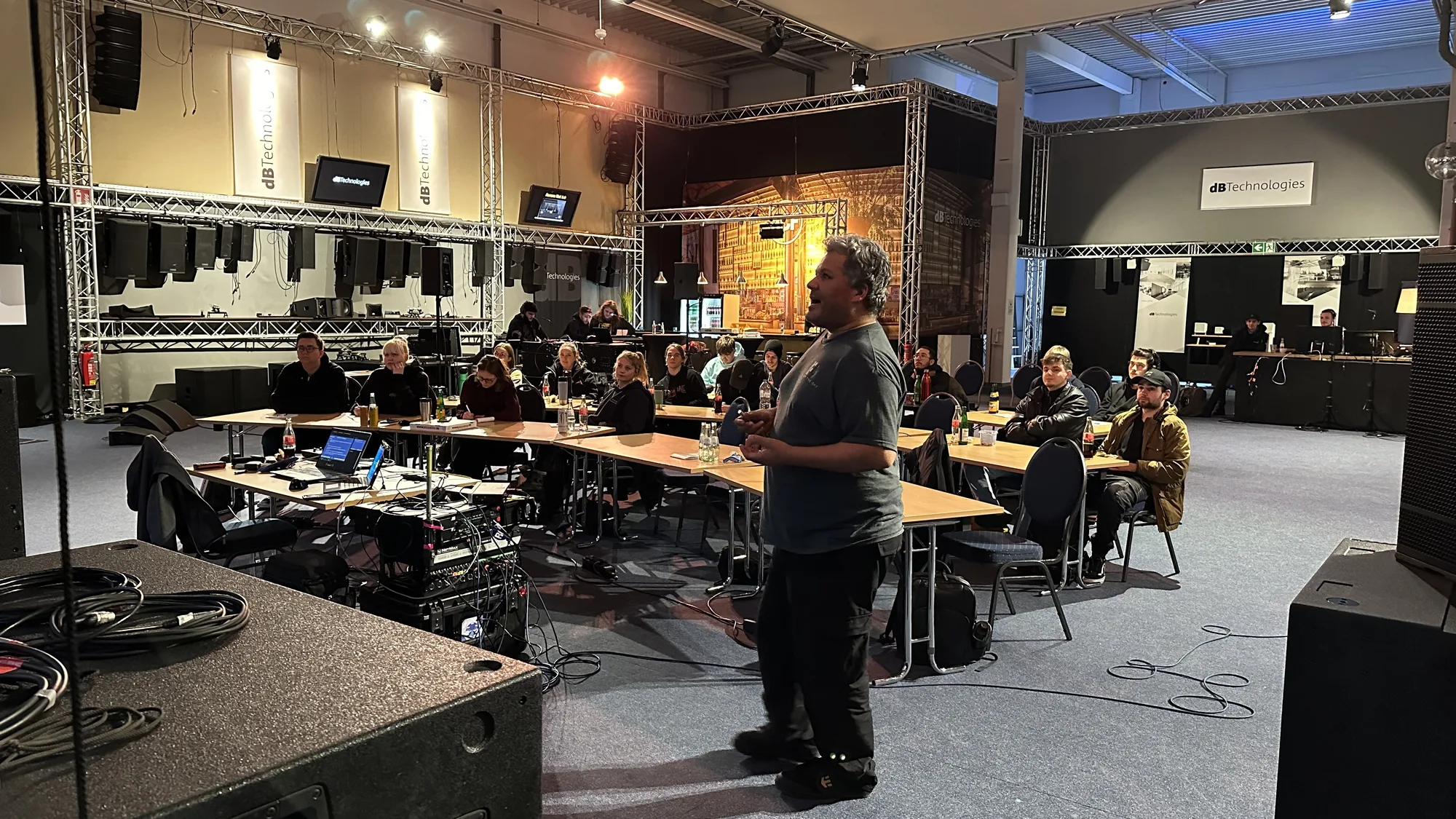 Seminarleiter erklärt vor einer Gruppe sitzender Teilnehmender in einer Halle mit Beschallungsanlage und Traversen